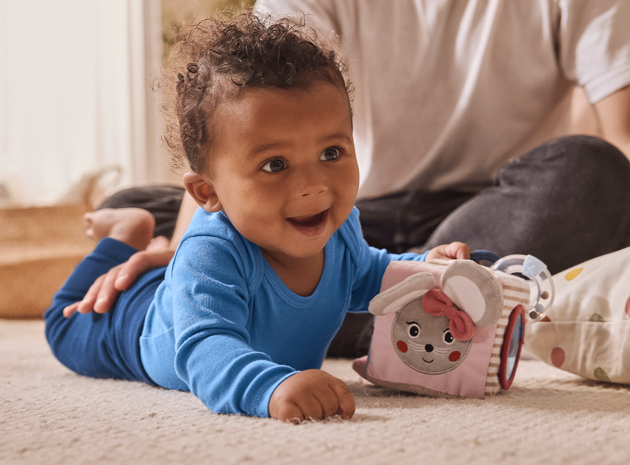 Bébé souriant en body bleu, jouant avec un cube d'activités en forme de souris.