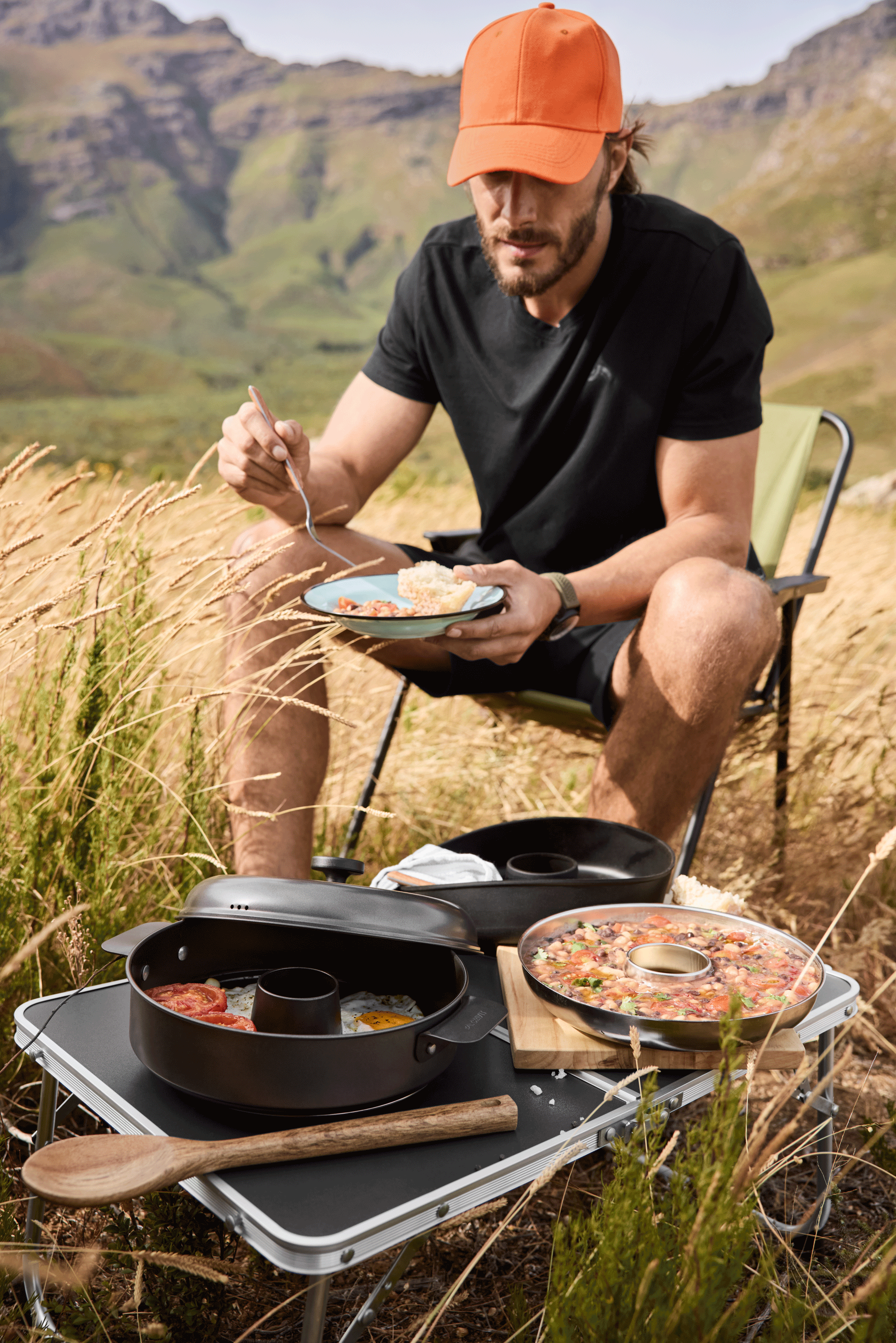 Homme en casquette orange mangeant un repas de camping avec des casseroles sur une table pliante.