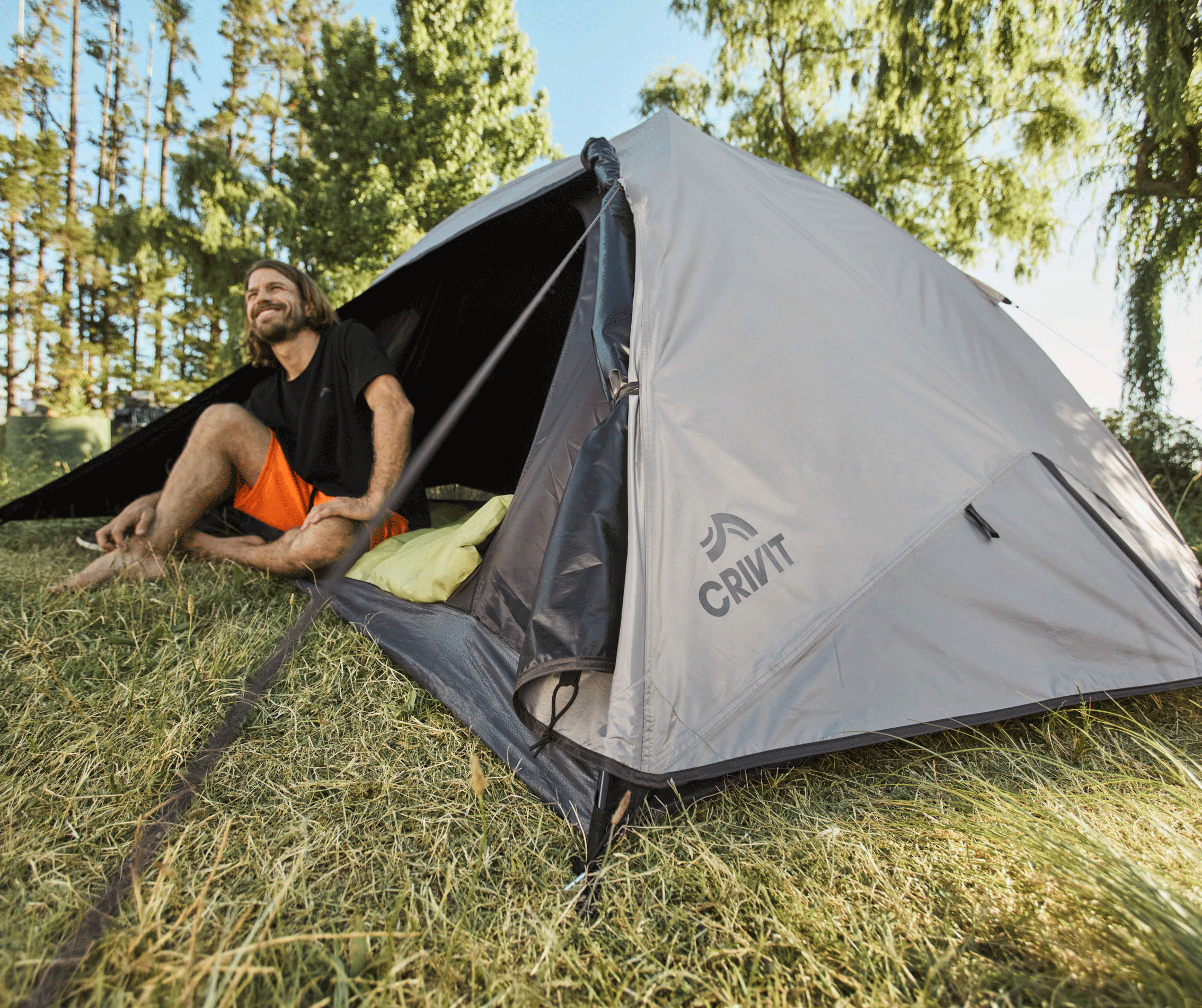 Un homme souriant est assis dans une tente de camping grise, entouré d'herbe et d'arbres.
