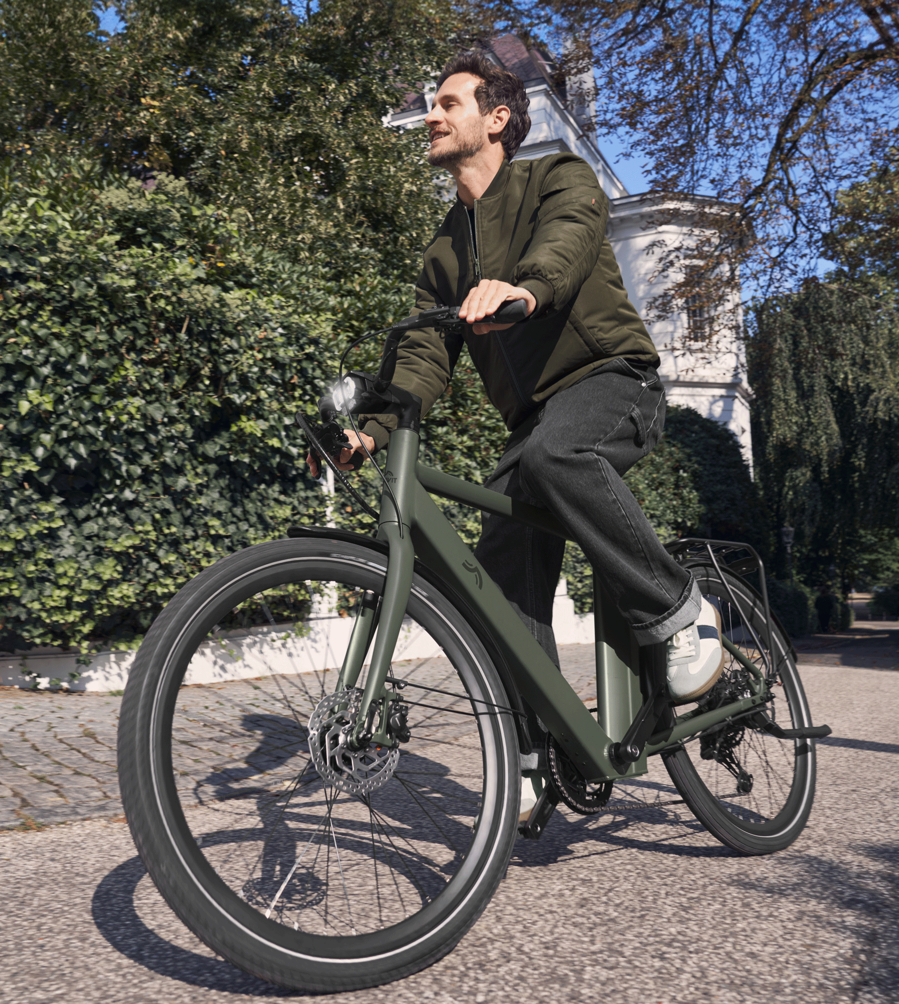 Homme souriant à vélo électrique vert sur une route ensoleillée avec des arbres et un bâtiment.