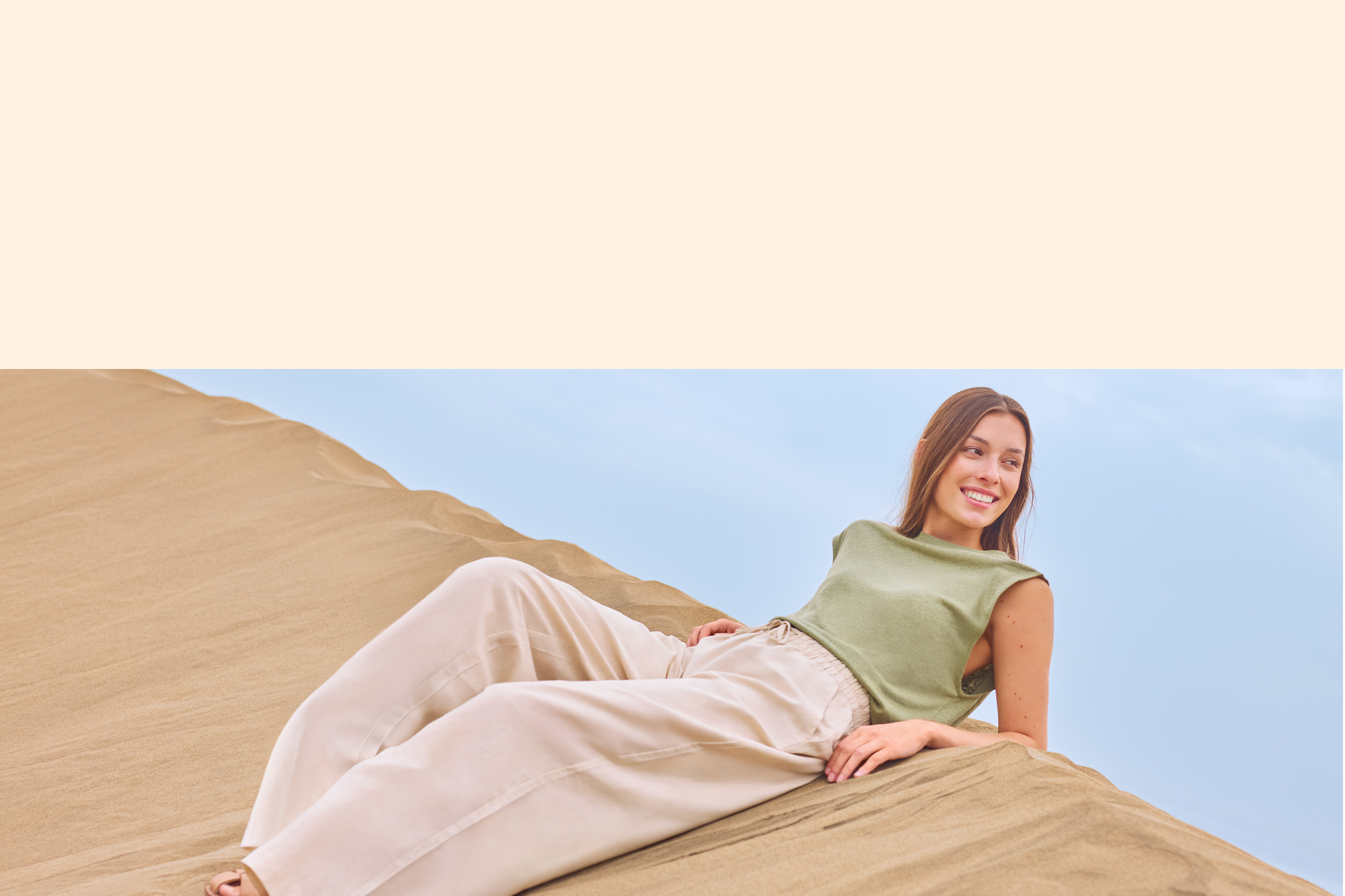 Femme souriante en haut vert et pantalon beige, allongée sur une dune de sable.