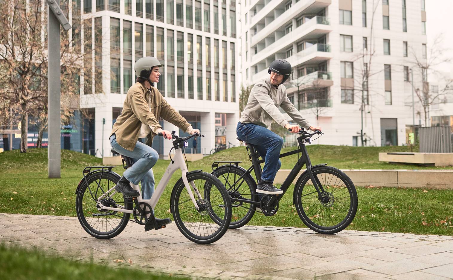 Deux personnes souriantes à vélo sur une piste cyclable, avec des bâtiments modernes en arrière-plan.