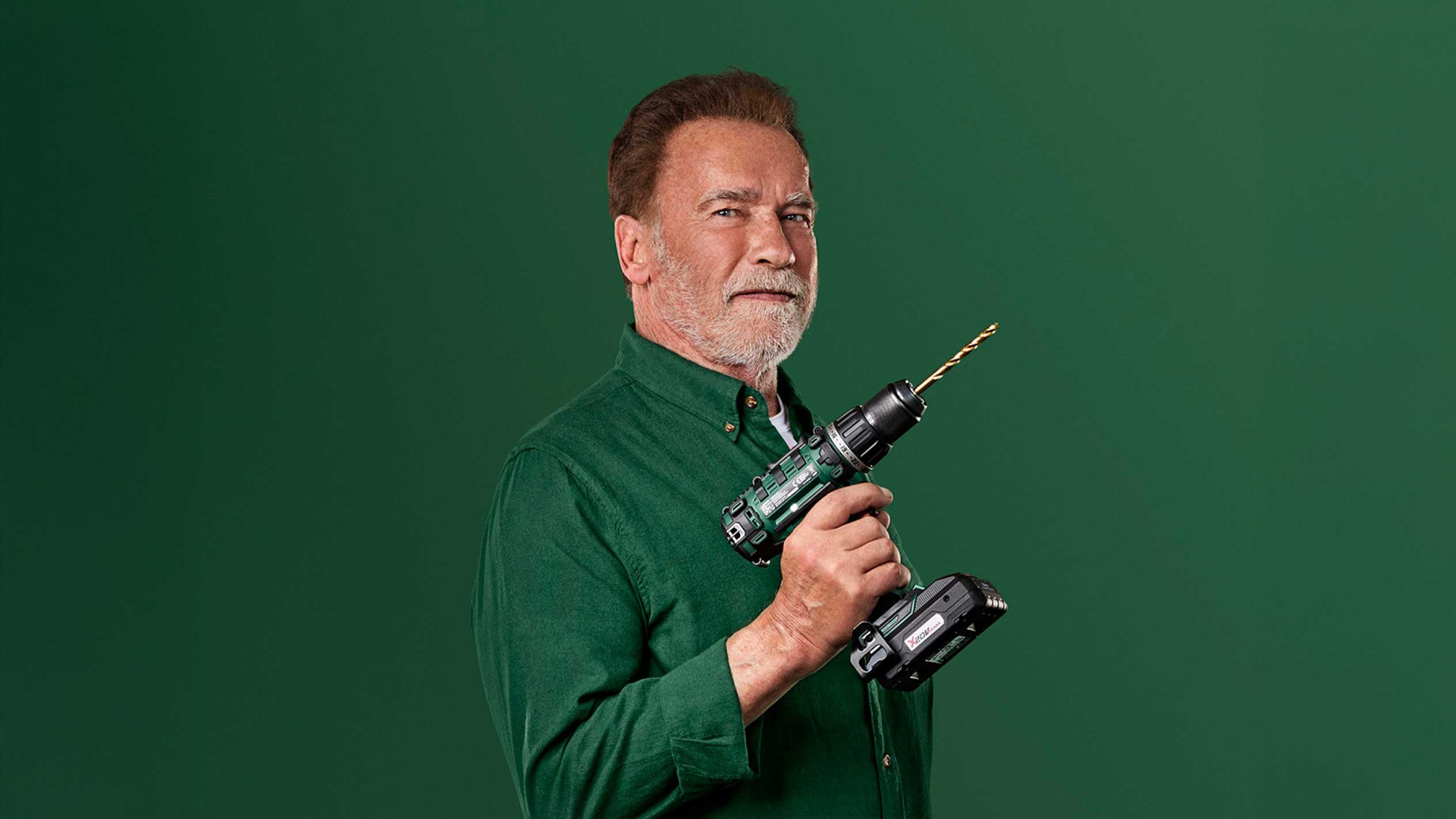 Arnold Schwarzenegger tenant une perceuse sans fil verte avec un foret doré.