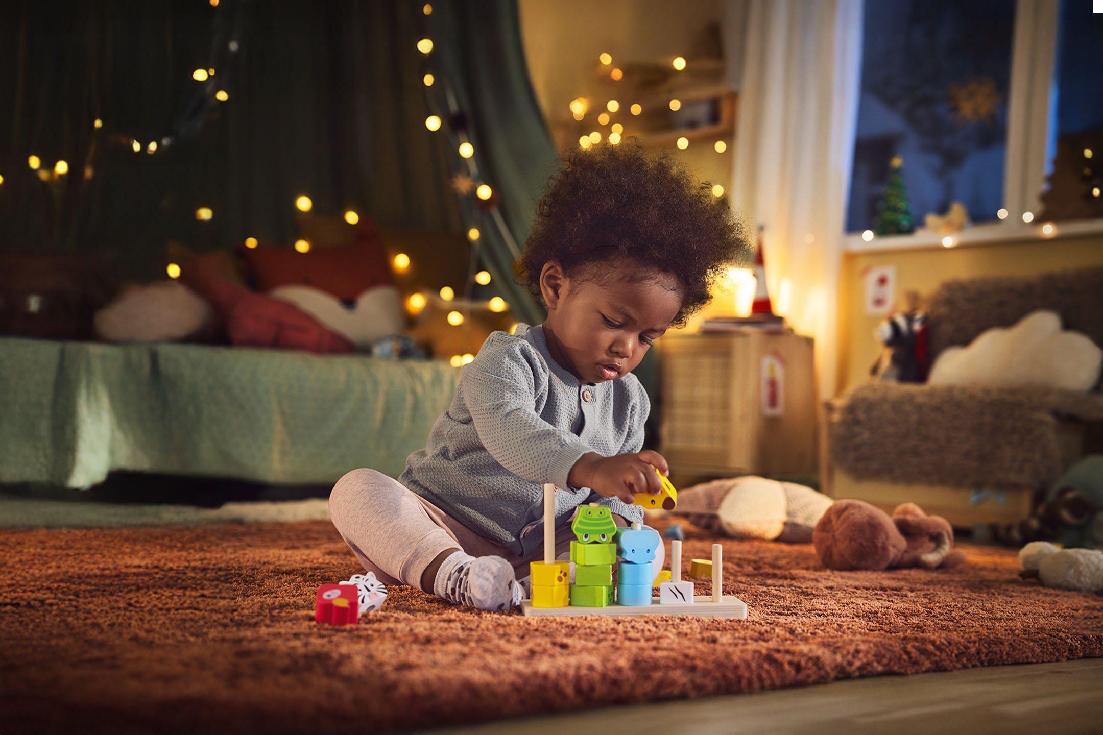 Bébé jouant avec un jouet d'empilement en bois sur un tapis moelleux, lumières de Noël en arrière-plan.