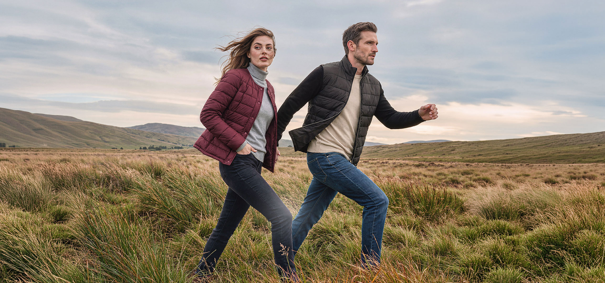 aiLabeling.aiLabelForImageAltText: Couple en doudounes légères et jeans marche dans un paysage vallonné et herbeux.