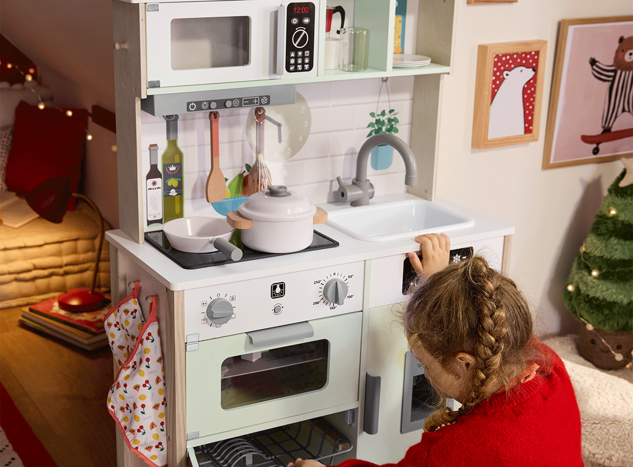 Une petite fille joue avec une cuisine en bois pour enfants, avec un four, un micro-ondes et un évier.
