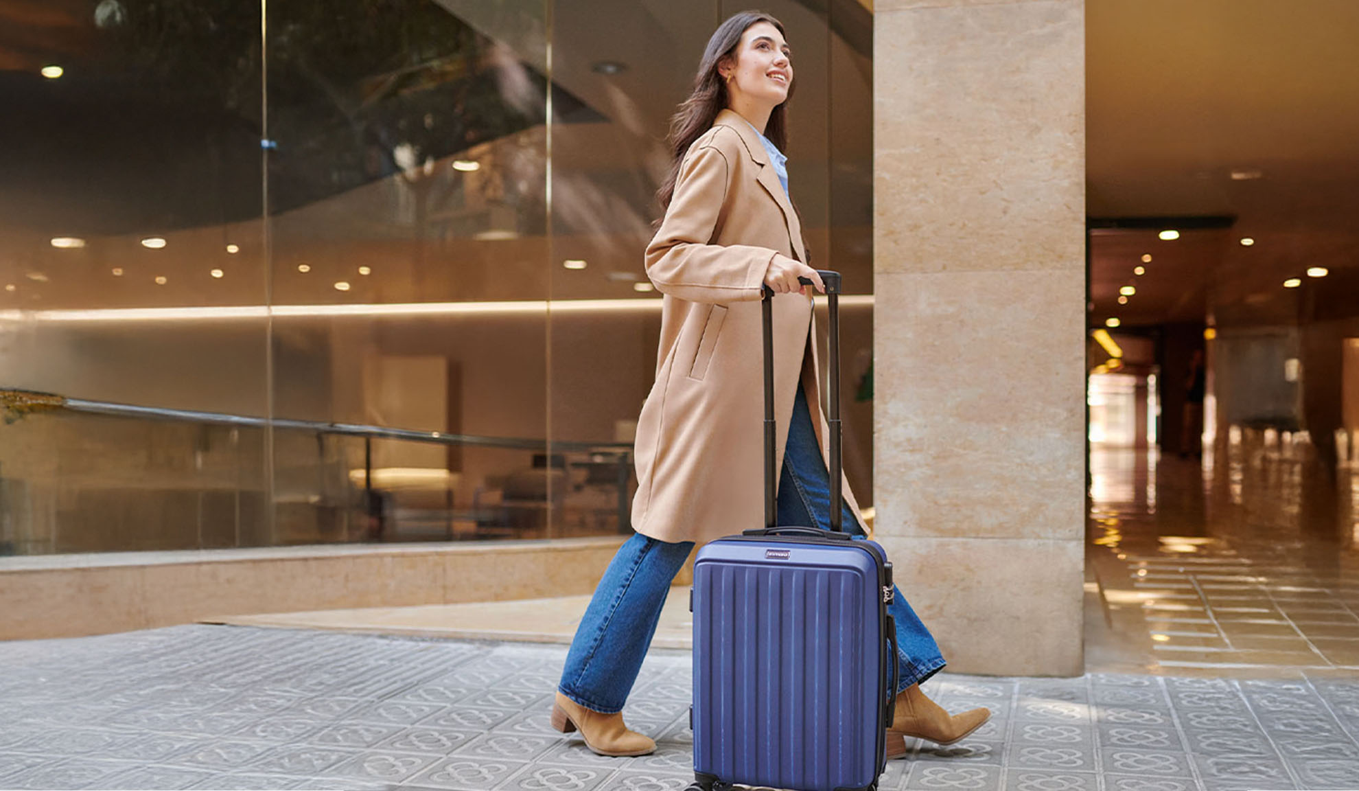 Femme en manteau beige et jean tirant une valise bleue sur un trottoir à motifs.