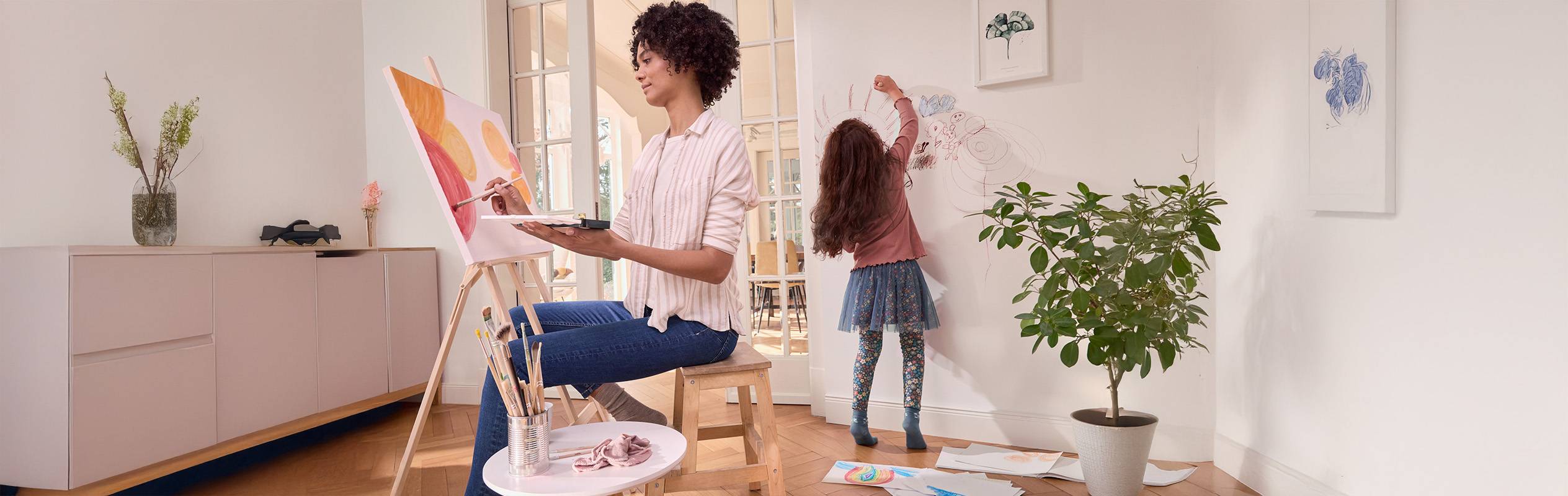 Une femme peint sur une toile tandis qu'une enfant dessine sur le mur à côté d'une plante.