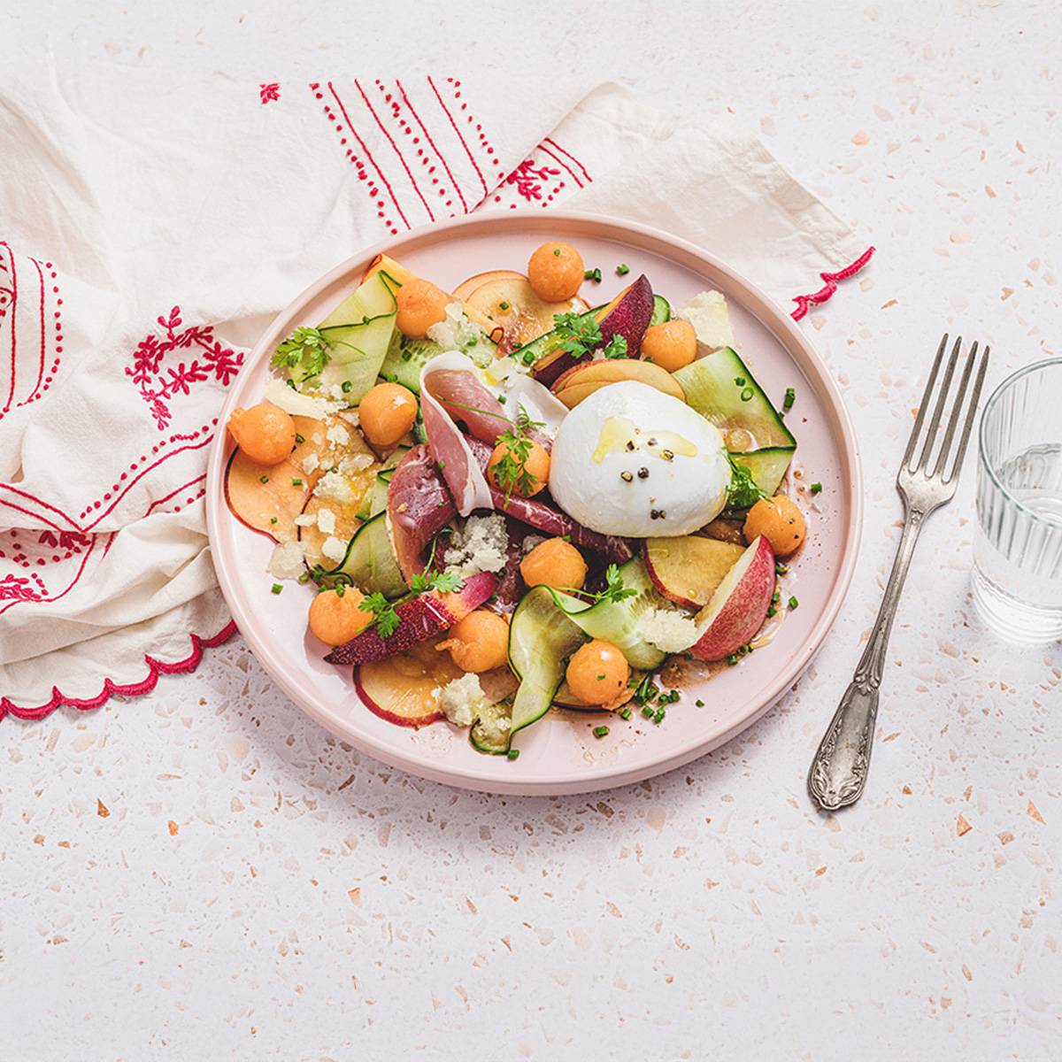 Salade estivale avec melon, prosciutto, burrata et concombre sur une table en terrazzo.
