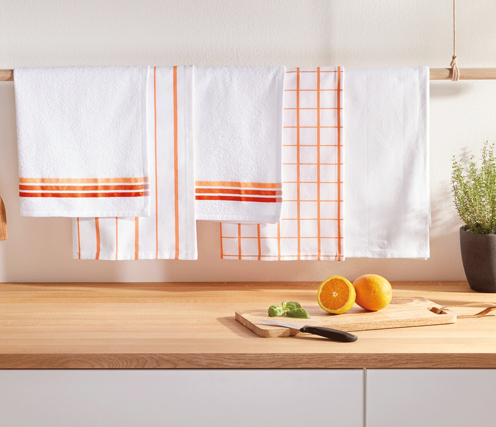 Torchons de cuisine blancs à rayures et carreaux orange, avec des oranges et une plante sur un comptoir en bois.