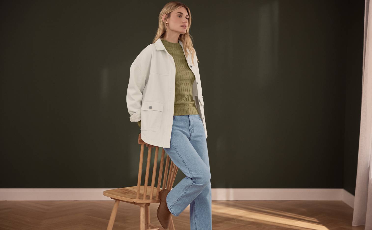 Femme en veste blanche, pull vert et jean bleu, posant sur une chaise en bois.