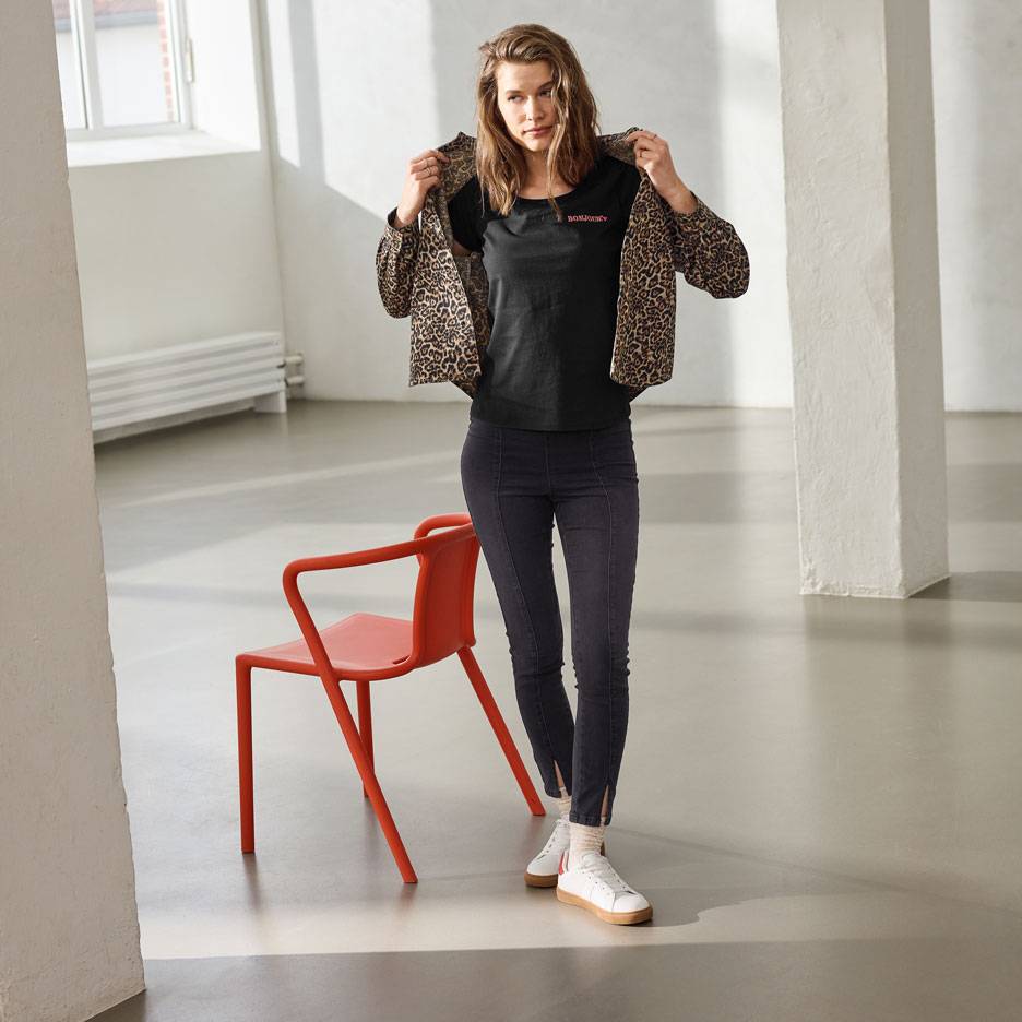 Femme en t-shirt noir avec texte, jean et veste léopard, debout à côté d'une chaise rouge.