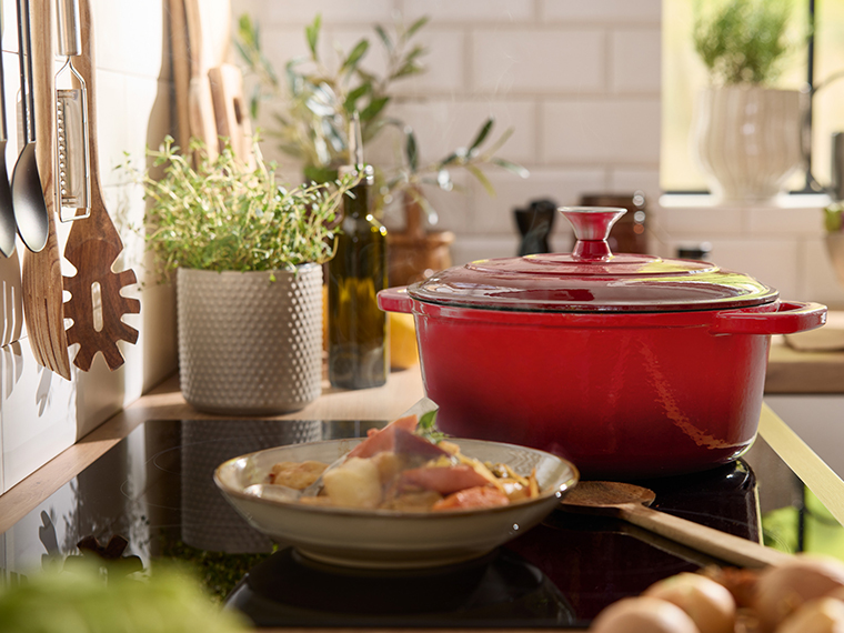 Une cocotte rouge sur une plaque de cuisson avec un plat de légumes à côté.