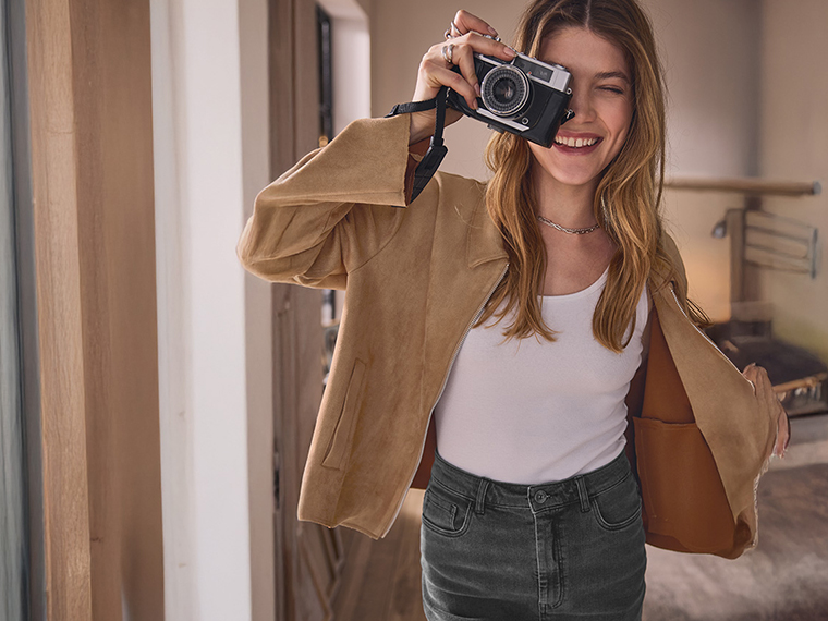 Une femme souriante en veste beige et jean gris tient un appareil photo vintage.