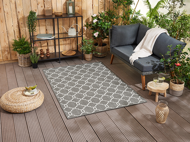Tapis d'extérieur gris à motif géométrique placé sur une terrasse en bois, près d'un canapé et de plantes.