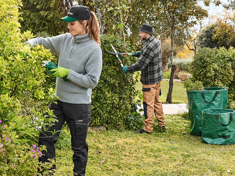 Deux personnes taillant des haies dans un jardin avec des outils de jardinage et des sacs de déchets verts.