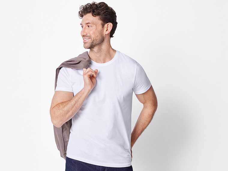 Un homme souriant en t-shirt blanc et jean, avec un pull sur l'épaule.