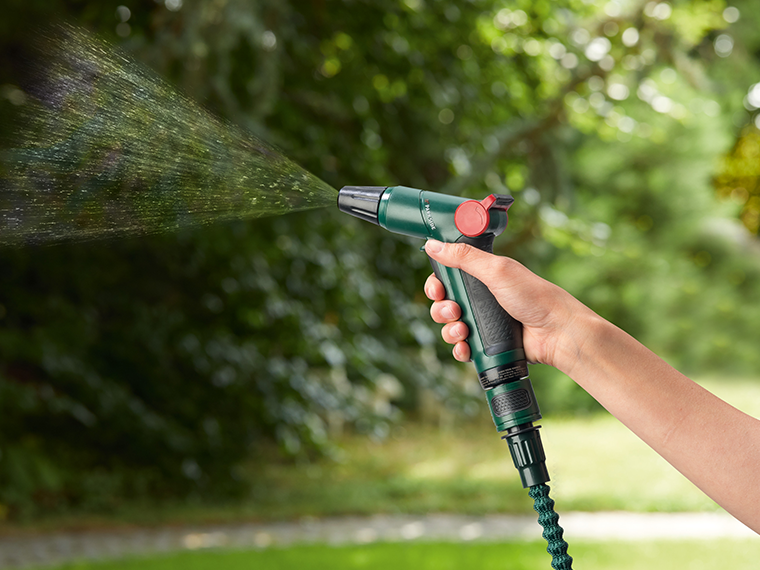 Une main tient un pistolet d'arrosage vert pulvérisant de l'eau dans un jardin.