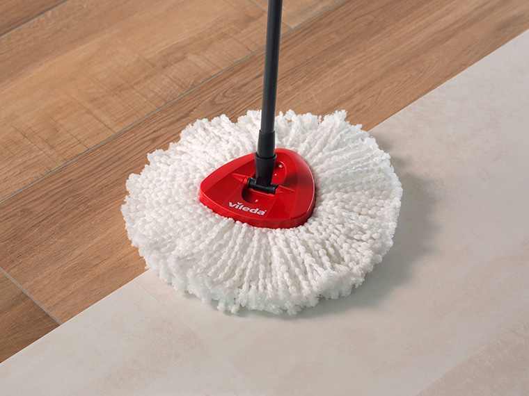 Un balai Vileda avec une tête de serpillière microfibre blanche nettoie un sol en bois.