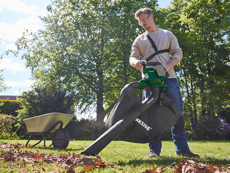 Un homme utilise un souffleur de feuilles Parkside pour ramasser les feuilles dans un jardin.