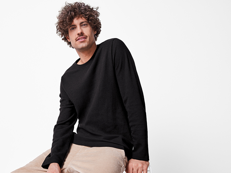 Un homme aux cheveux bouclés porte un pull noir et un pantalon beige.