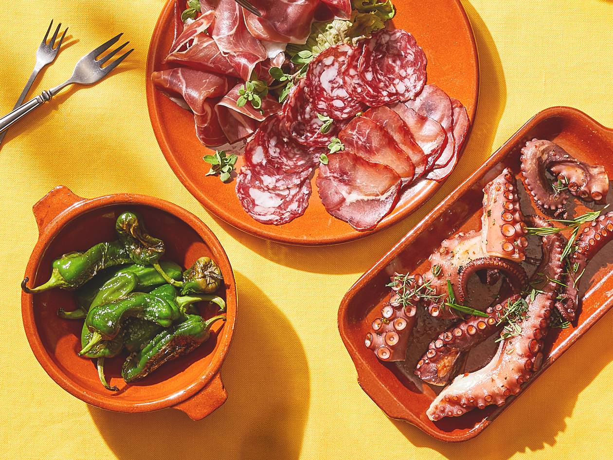 Un assortiment de tapas espagnoles avec de la charcuterie, du poulpe et des piments padrón sur une nappe jaune.