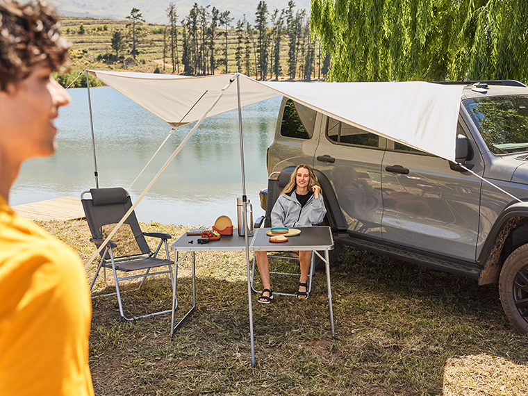 Un couple campe au bord d'un lac avec un auvent de voiture, une table et des chaises de camping.
