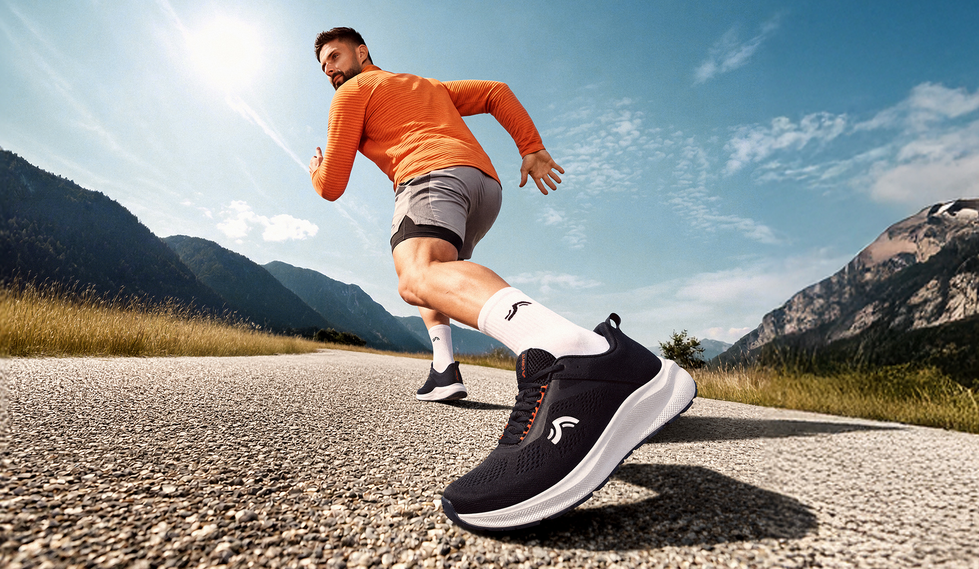 Homme en tenue de sport orange et grise, courant sur une route de montagne ensoleillée.