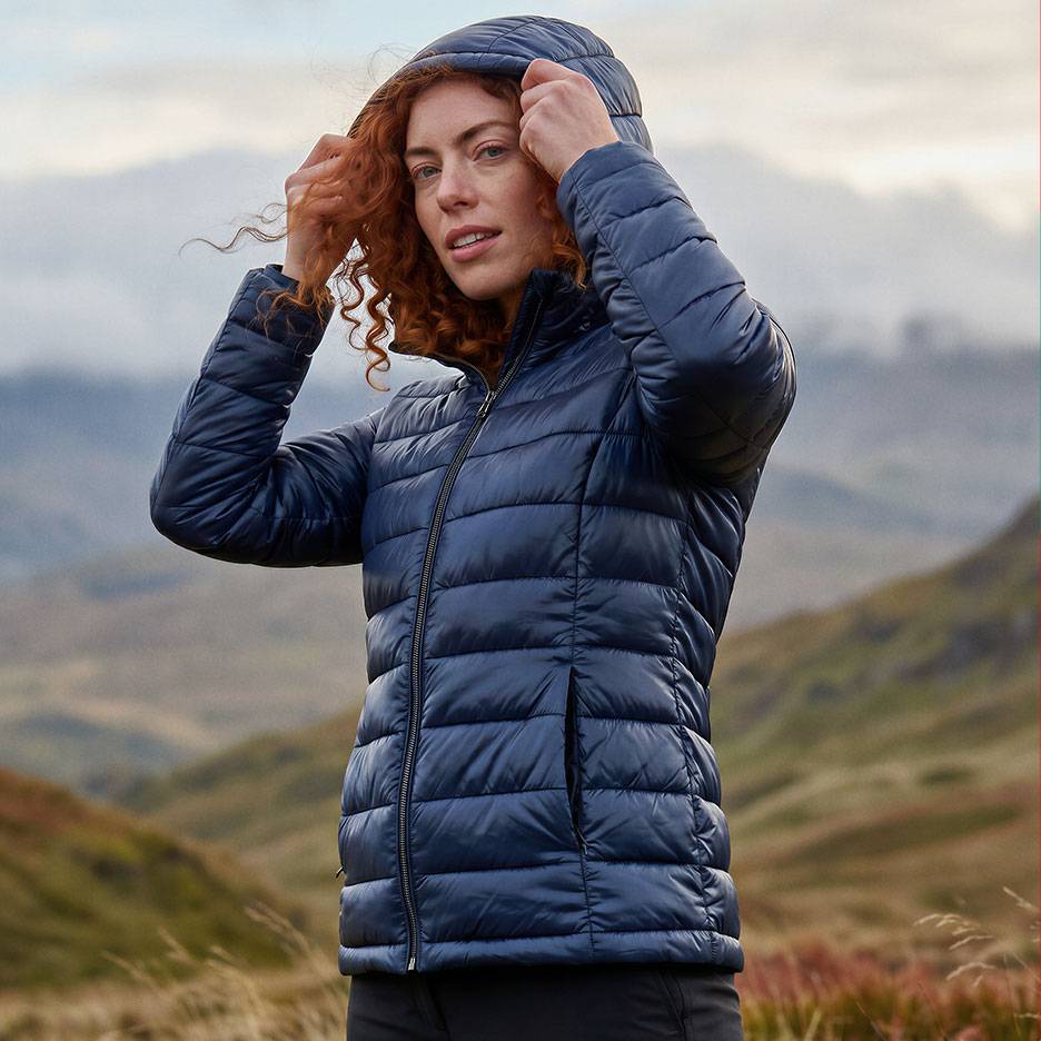 Femme aux cheveux roux dans une doudoune bleue foncée à capuche dans un paysage montagneux.