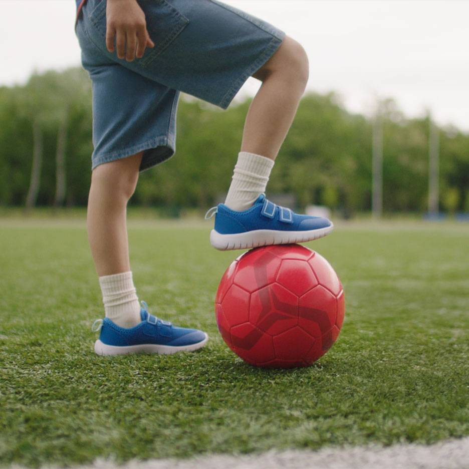 Enfant en short et baskets bleues, avec un pied sur un ballon de football rouge.