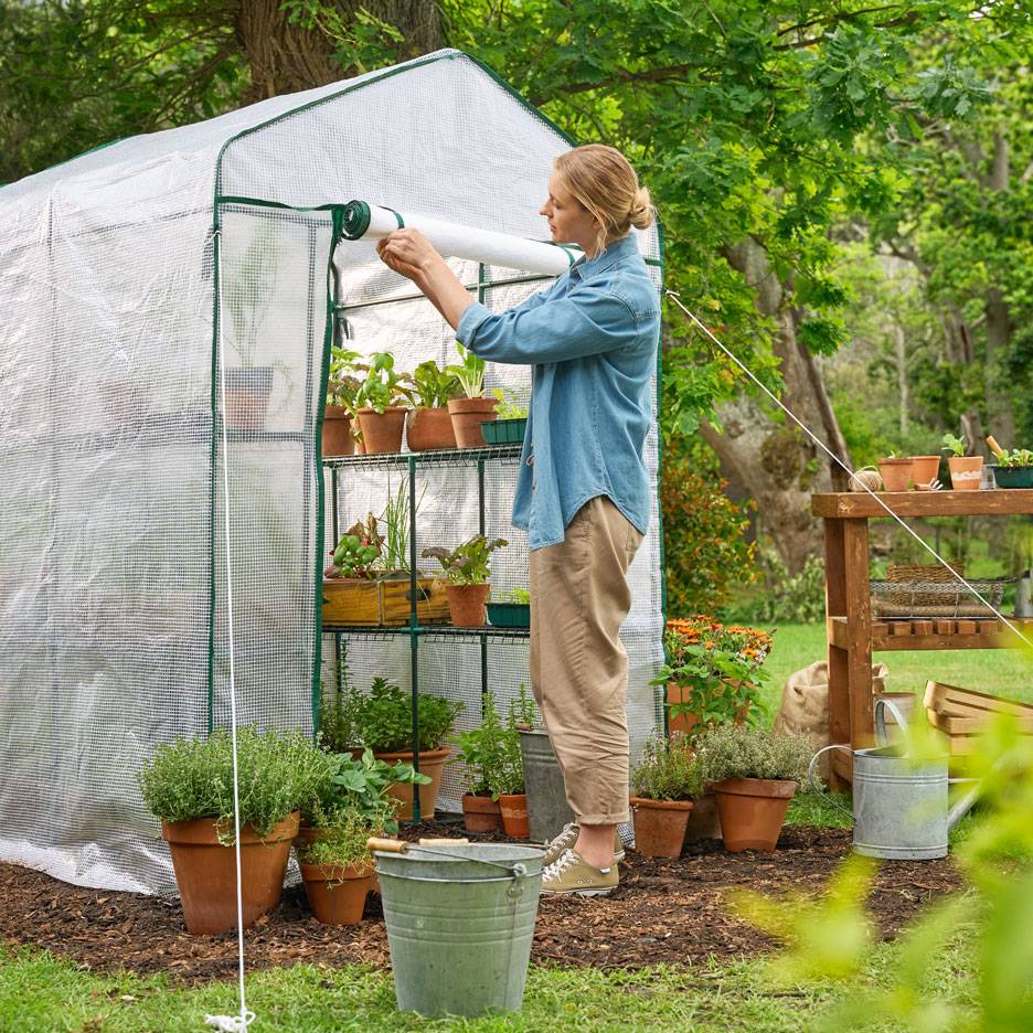 Femme s'occupant de plantes dans une serre avec des étagères d'herbes et de légumes en pot.