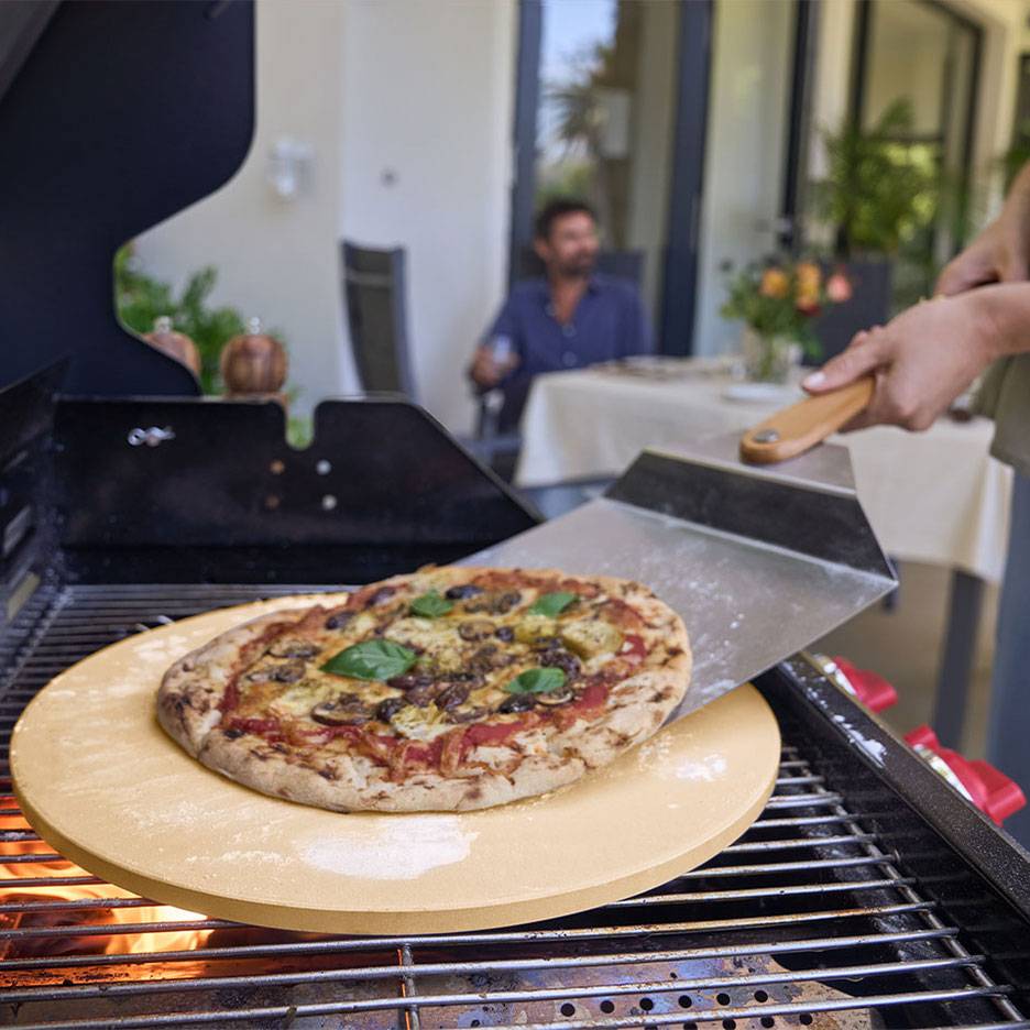 Pizza sur une pierre à pizza sur un grill, avec une pelle à pizza et un homme en arrière-plan.