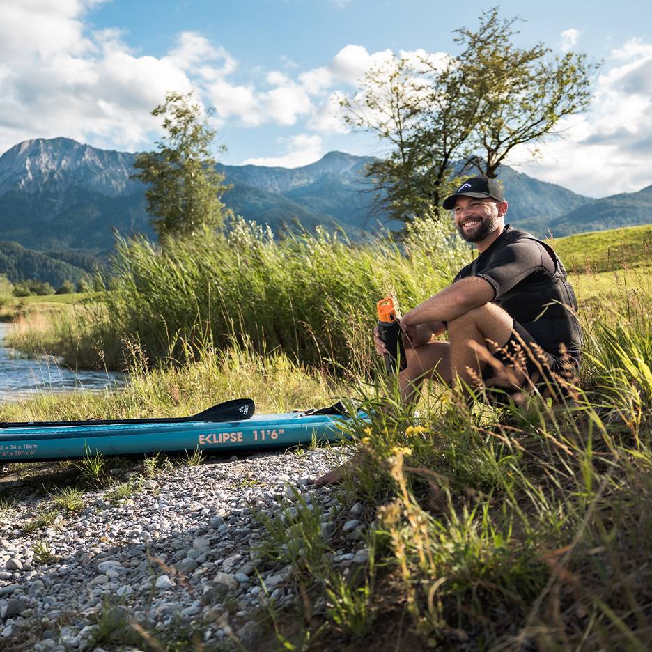 Homme souriant avec casquette et gilet de sauvetage, assis près d'une planche de paddleboard bleue au bord de la rivière.