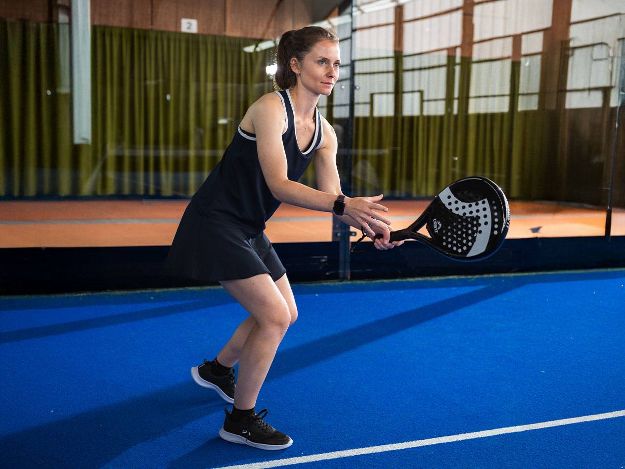 Femme en robe de padel noire avec raquette de padel noire sur terrain bleu.