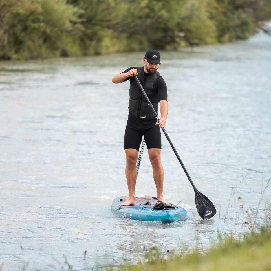 Homme faisant du paddleboard sur une rivière, portant une combinaison, un gilet de sauvetage et une casquette noirs.