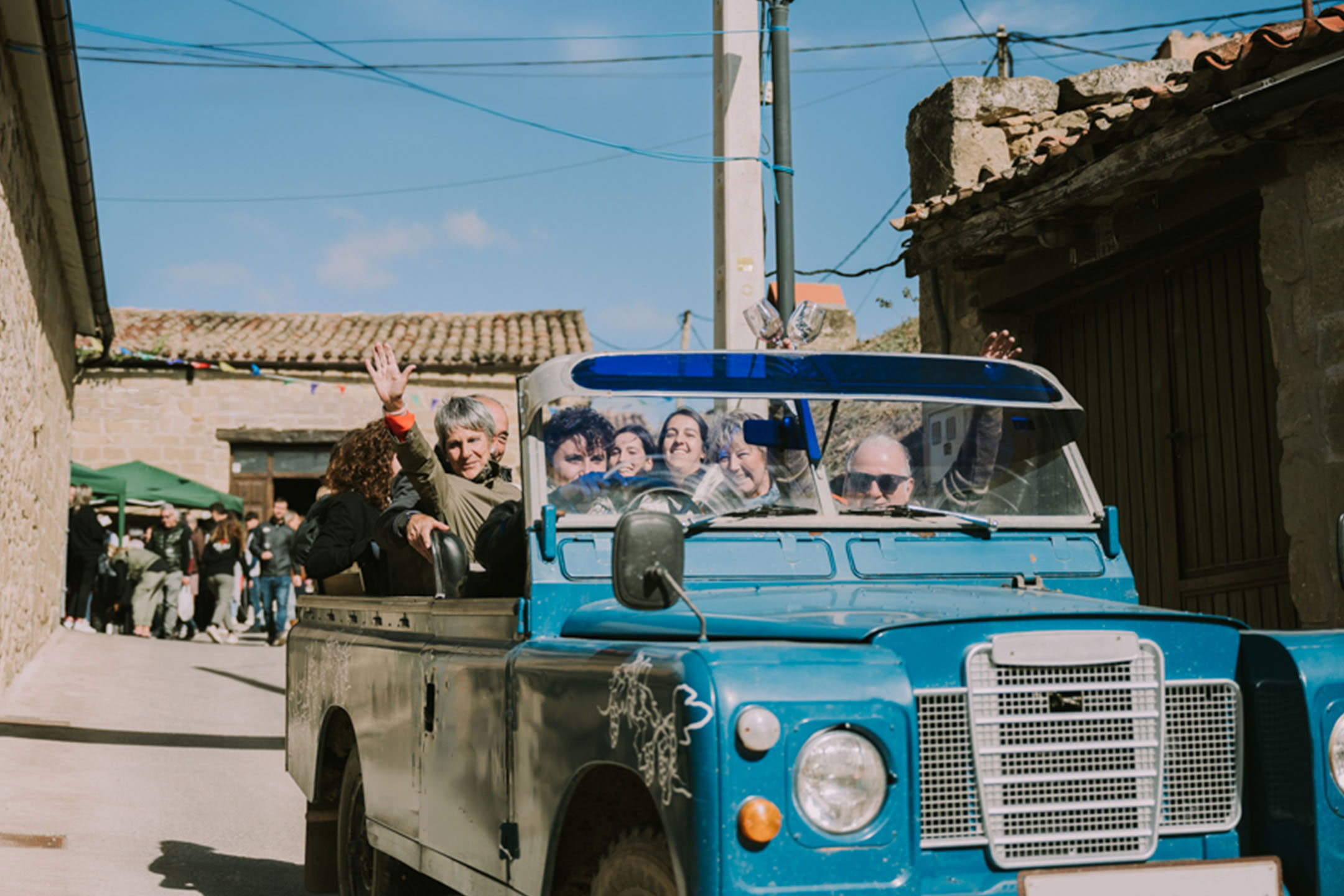 Des personnes dans un véhicule tout-terrain vintage bleu traversant une rue de village.