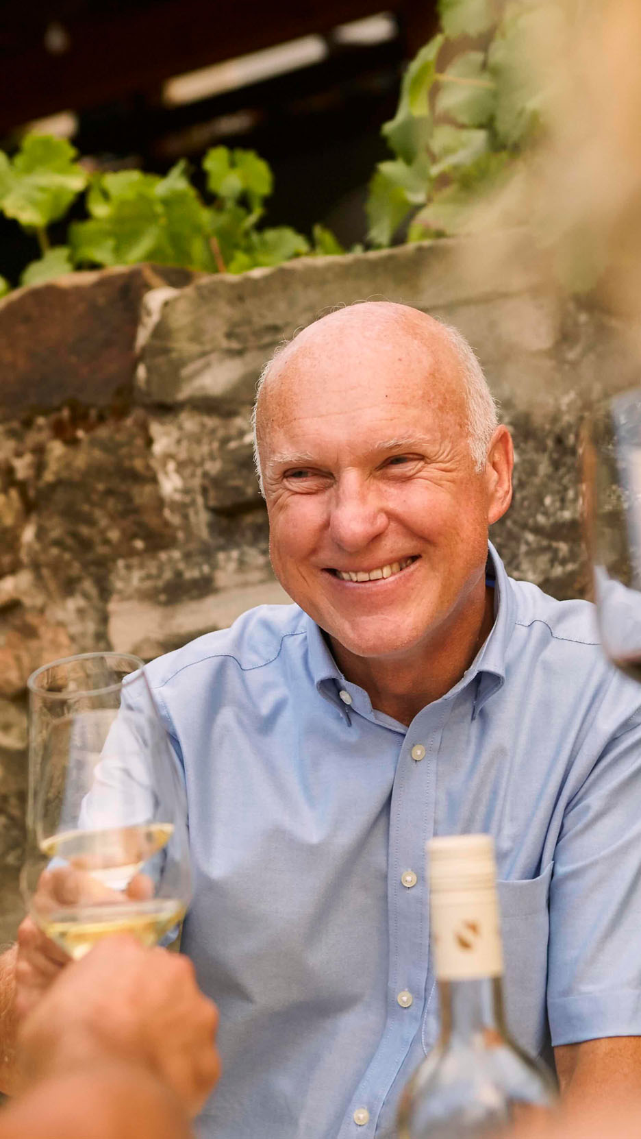 Homme souriant en chemise bleue trinquant avec un verre de vin blanc en extérieur.