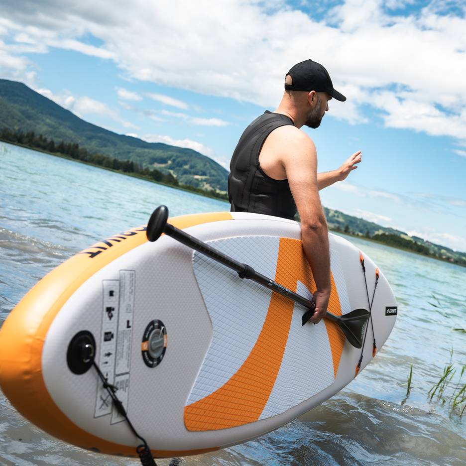 Homme en gilet de sauvetage et casquette tenant une planche de paddle gonflable dans un lac.