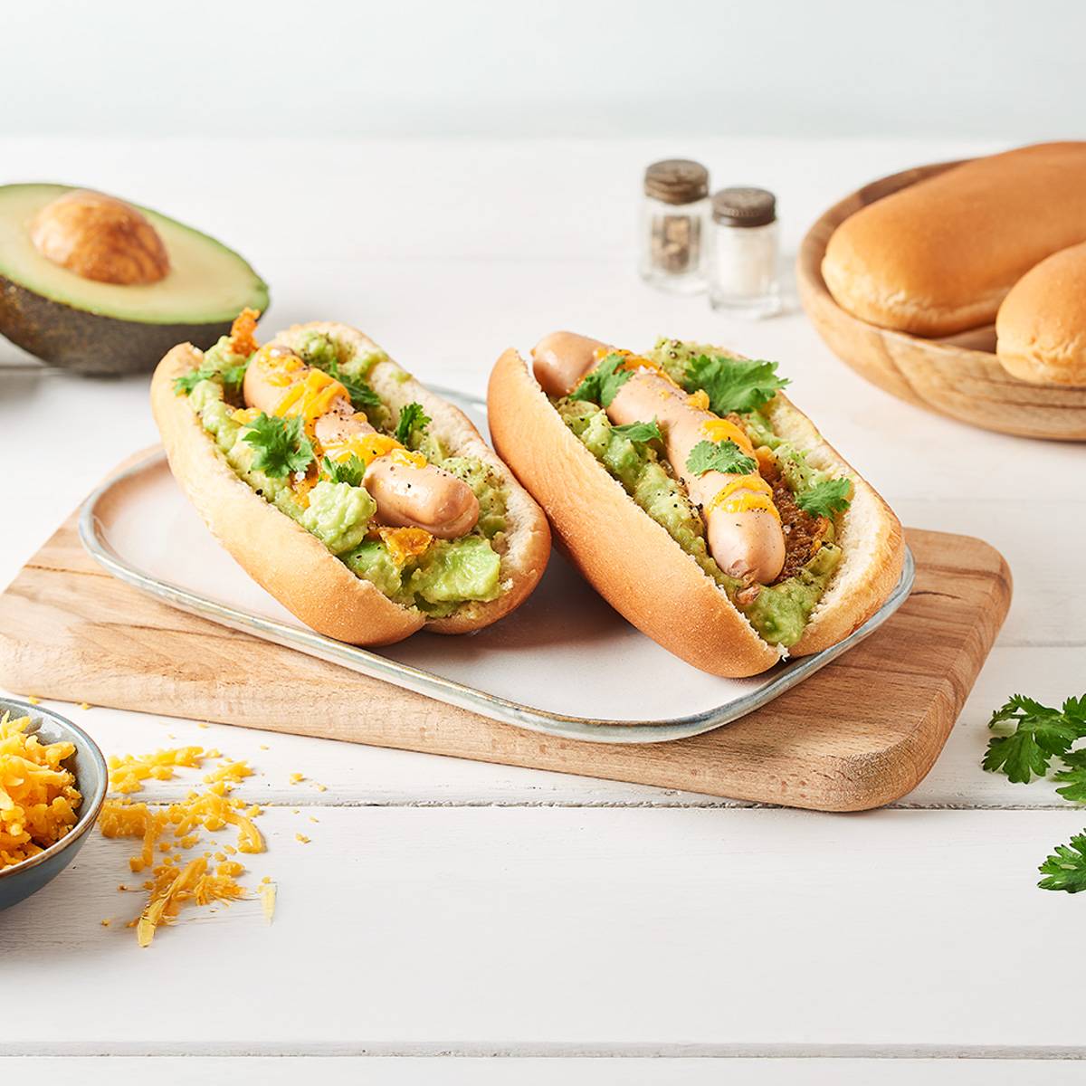 Deux hot-dogs à l'avocat et au fromage sur une planche en bois, avec des pains et un avocat en arrière-plan.
