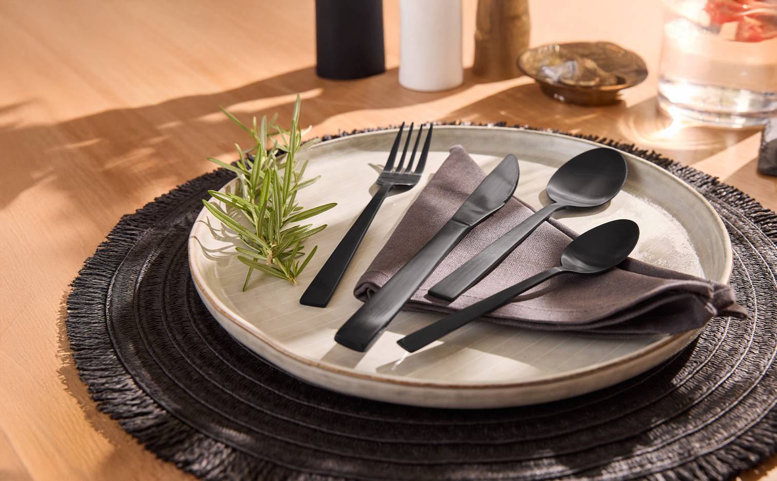 Couverts noirs mats sur une assiette en céramique avec une serviette et du romarin.