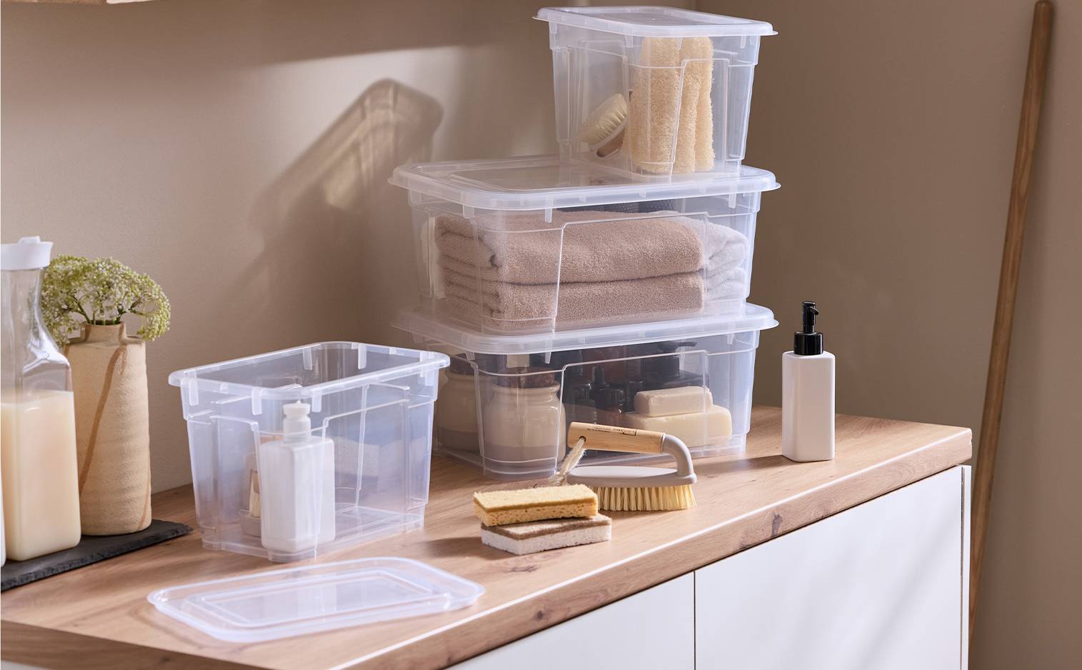 Boîtes de rangement transparentes empilées avec serviettes, brosses et savons sur un comptoir en bois.