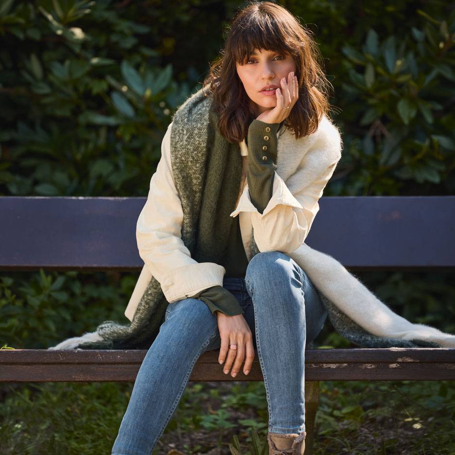 Femme en jean, veste blanche et écharpe verte assise sur un banc de parc.