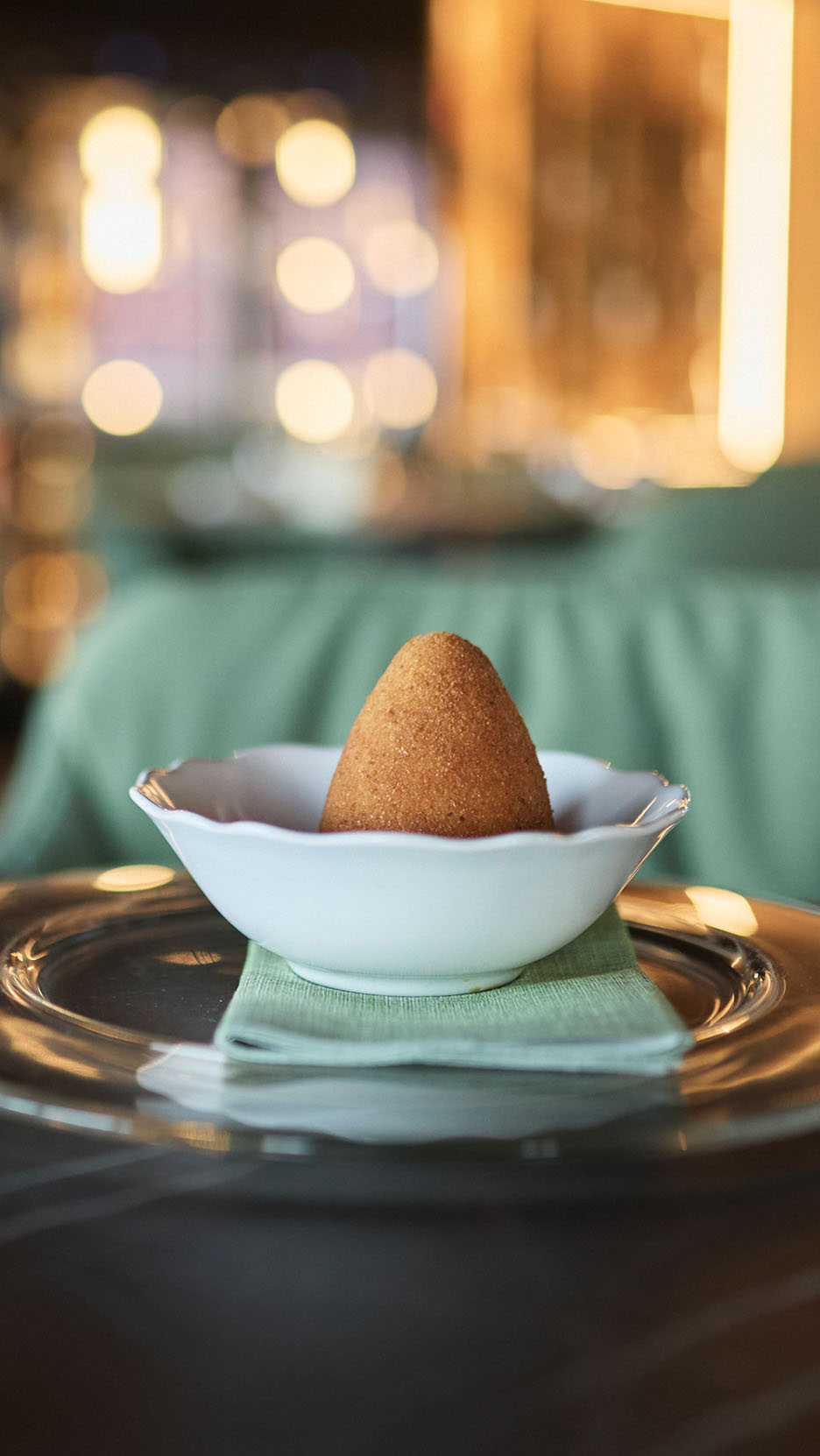 Arancini doré dans un bol blanc sur une nappe verte et une assiette en verre.