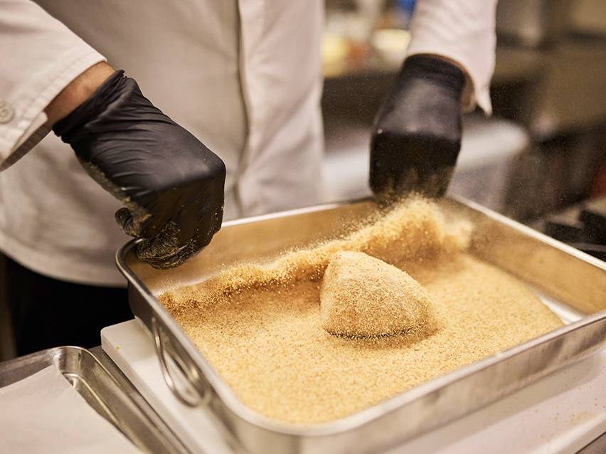 Un chef en gants noirs panant un aliment dans un plat en métal.