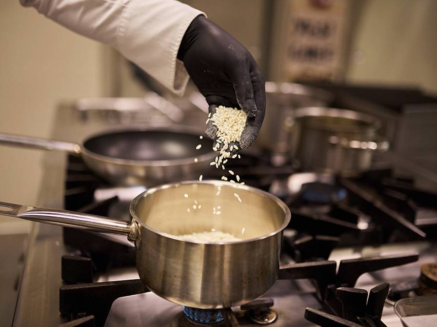 Un chef en gants noirs verse du riz dans une casserole sur une cuisinière à gaz.