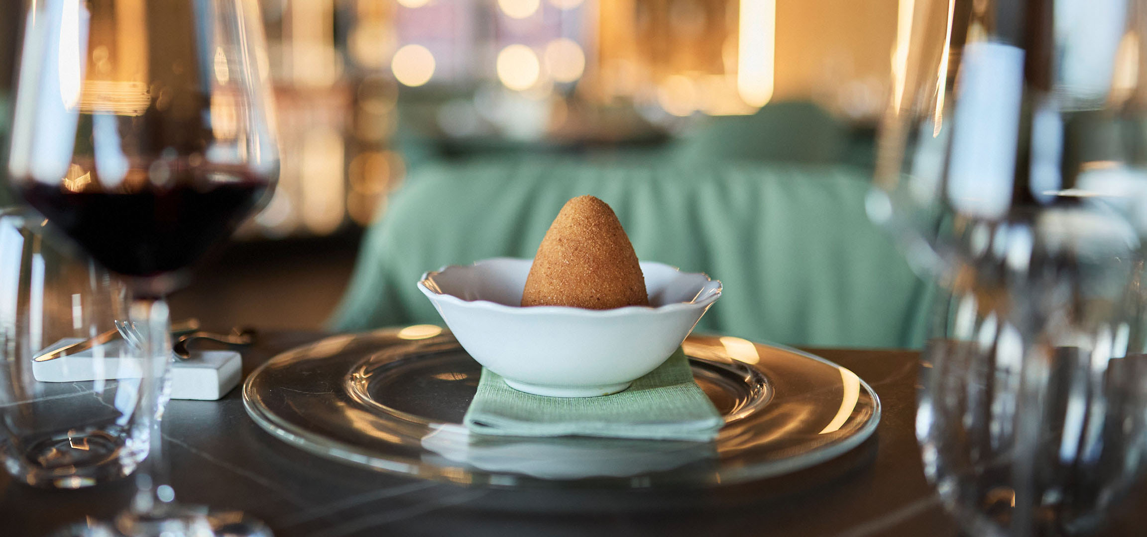 Un plat conique doré dans un bol blanc, avec du vin rouge et des couverts sur une table.