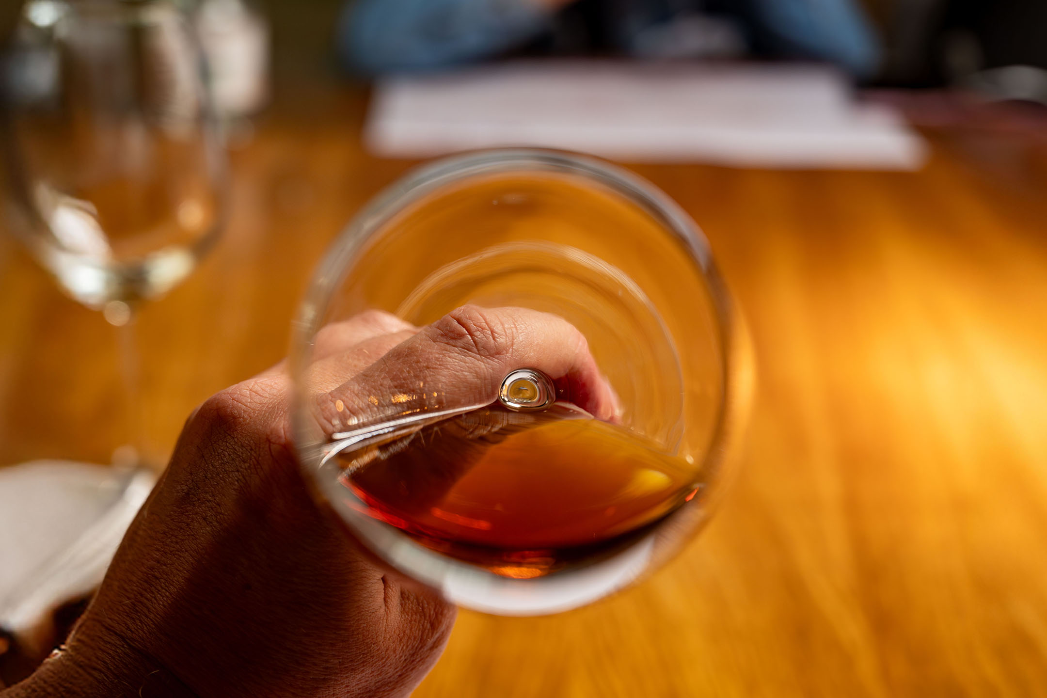 Main tenant un verre de liquide ambré, peut-être du brandy ou du whisky, sur une table en bois.