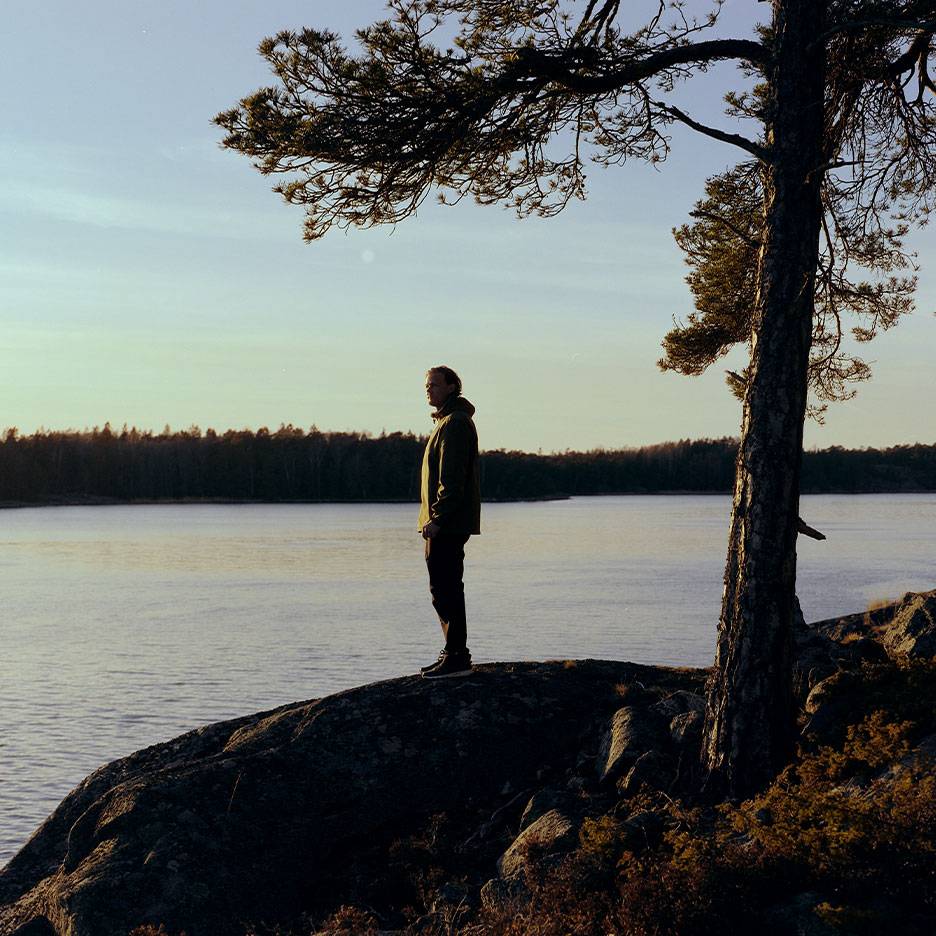 Homme en veste de randonnée au bord d'un lac, avec un arbre au premier plan.