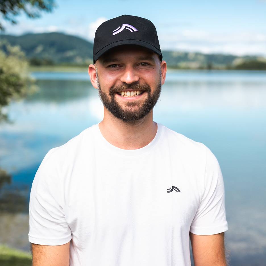 Homme souriant avec casquette et t-shirt blancs, avec lac et montagnes en arrière-plan.