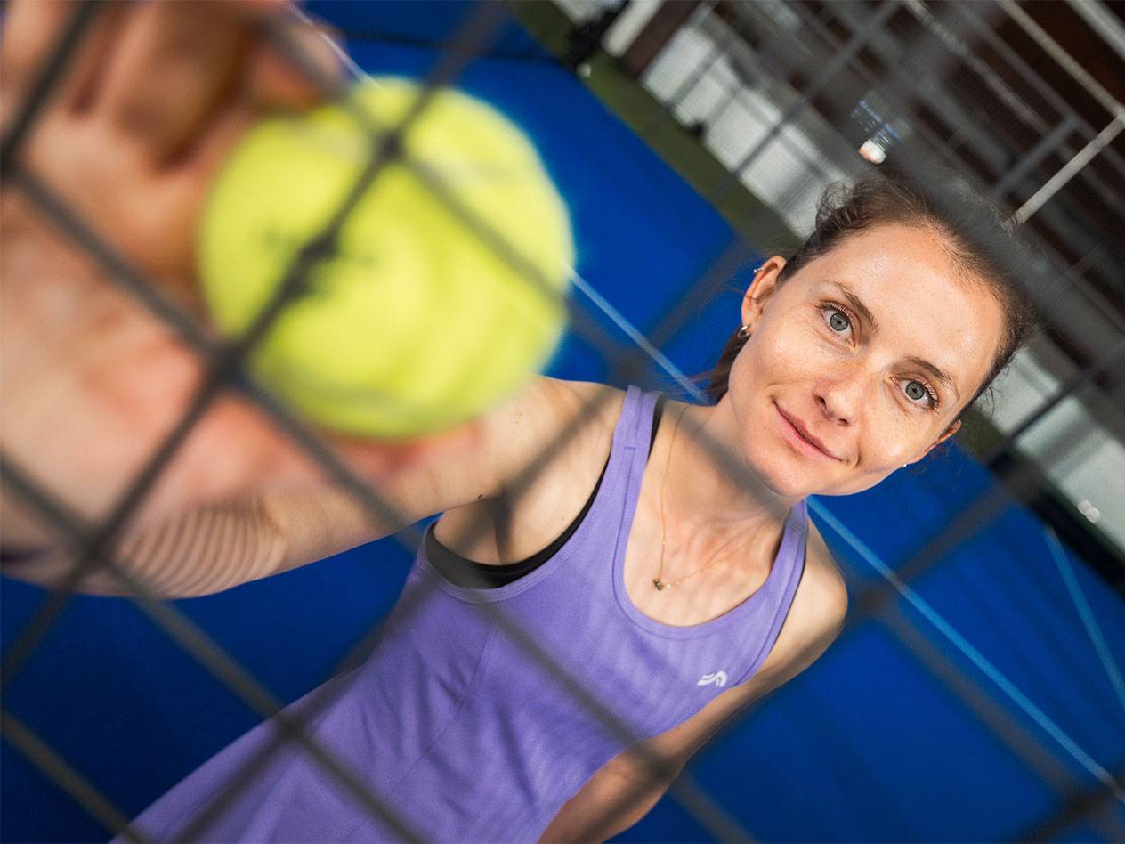 Femme en tenue de sport tenant une balle de padel à travers un filet, terrain bleu en arrière-plan.